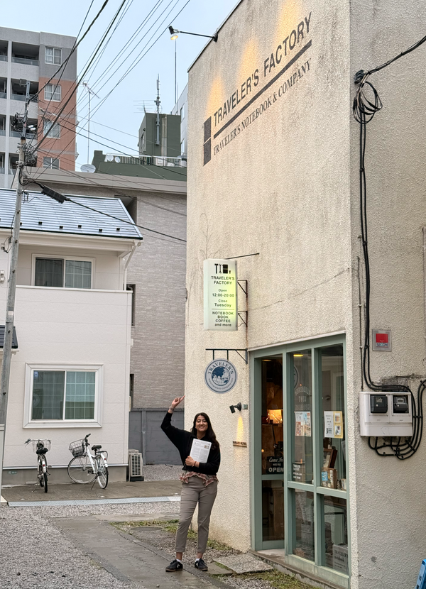 A woman stands in front of a building with a large sign reading 'Traveler's Factory' and 'Travelers' Neighbor & Company,' holding a small menu or sign. The exterior wall is textured beige, with a green-framed glass entrance and visible utility wiring.