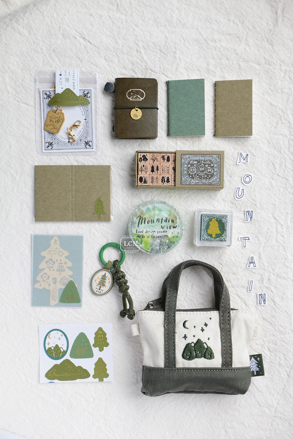 A styled flat-lay of a mountain-themed stationery and accessory set on a textured white fabric background. The set includes a canvas tote bag with a green and cream mountain graphic, a small notebook, a green journal, kraft paper envelopes, stickers, a keychain, and a circular 'Mountain Love' pin.