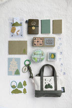 A styled flat-lay of a mountain-themed stationery and accessory set on a textured white fabric background. The set includes a canvas tote bag with a green and cream mountain graphic, a small notebook, a green journal, kraft paper envelopes, stickers, a keychain, and a circular 'Mountain Love' pin.