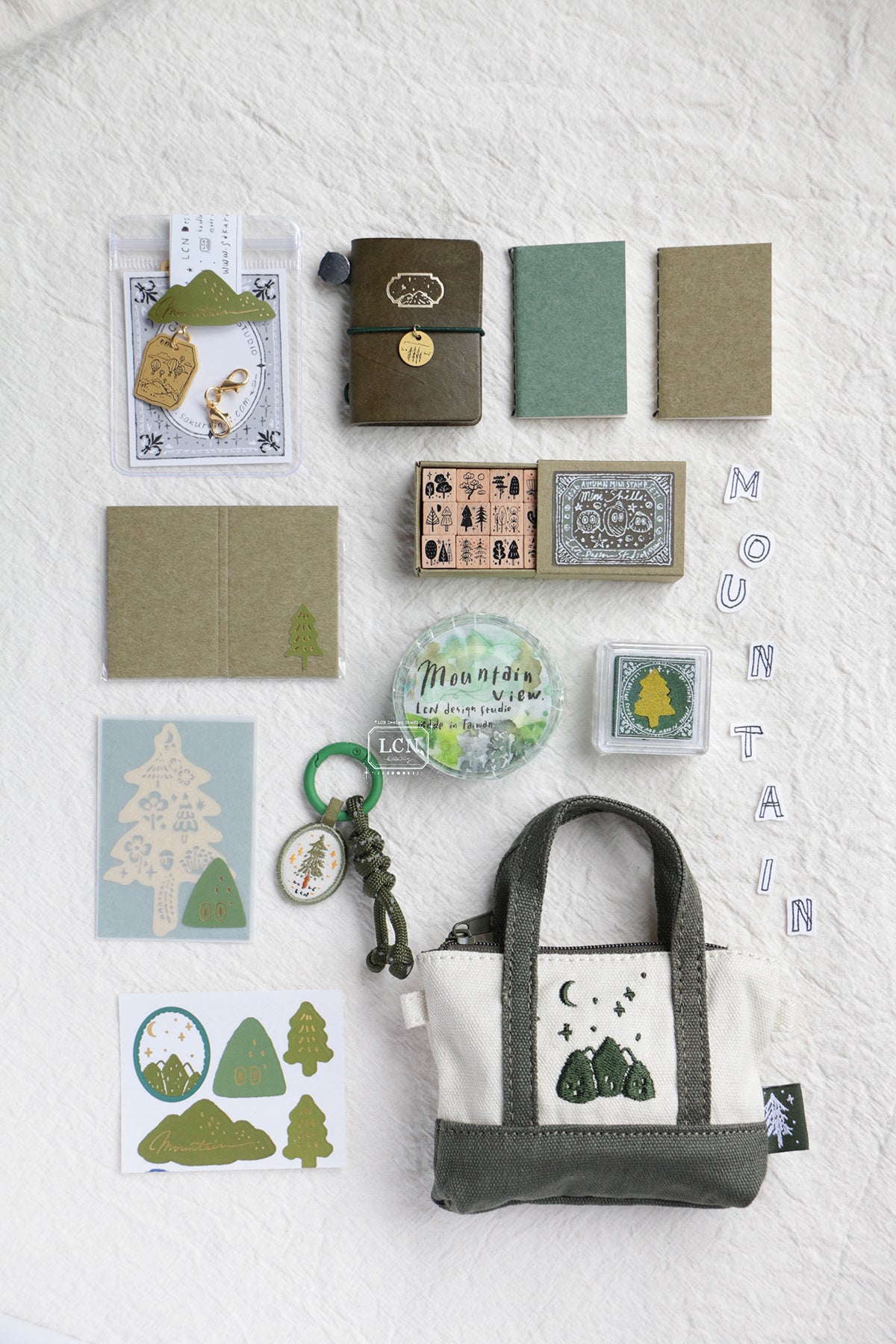 A styled flat-lay of a mountain-themed stationery and accessory set on a textured white fabric background. The set includes a canvas tote bag with a green and cream mountain graphic, a small notebook, a green journal, kraft paper envelopes, stickers, a keychain, and a circular 'Mountain Love' pin.