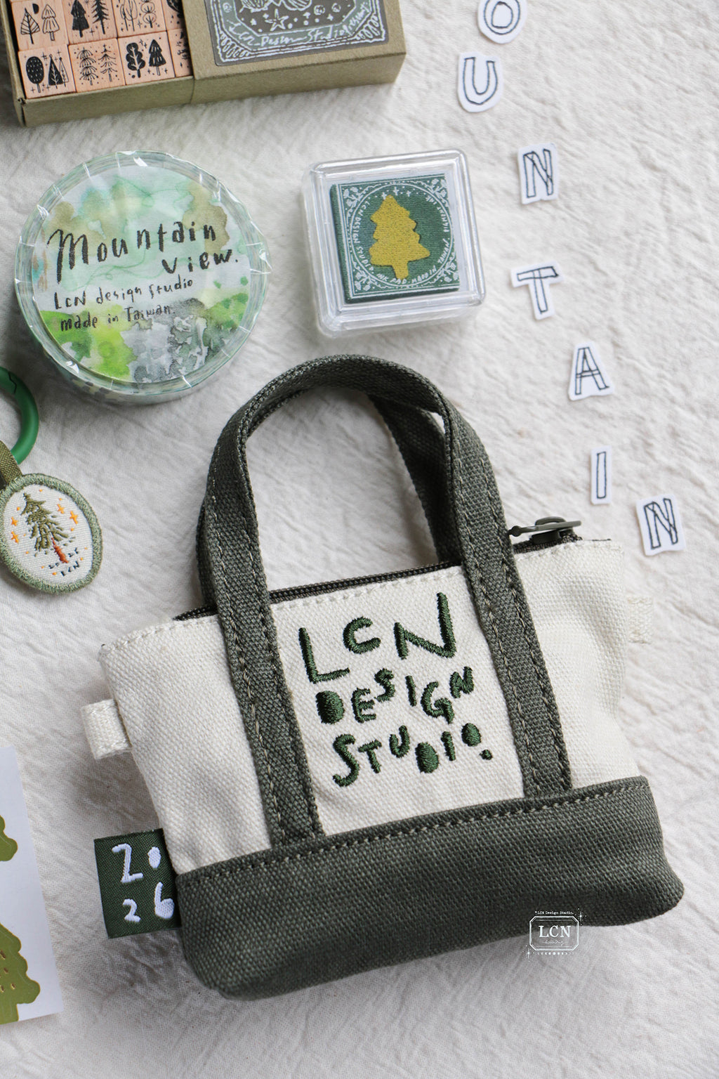 A small canvas tote-style pouch with dark green handles and base, and an off-white center panel reading 'LEN DESIGN STUDIO,' arranged on a textured white surface with craft-themed props including rubber stamps, tape, and letter tiles spelling MOUNTAIN.