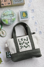 A small canvas tote-style pouch with dark green handles and base, and an off-white center panel reading 'LEN DESIGN STUDIO,' arranged on a textured white surface with craft-themed props including rubber stamps, tape, and letter tiles spelling MOUNTAIN.